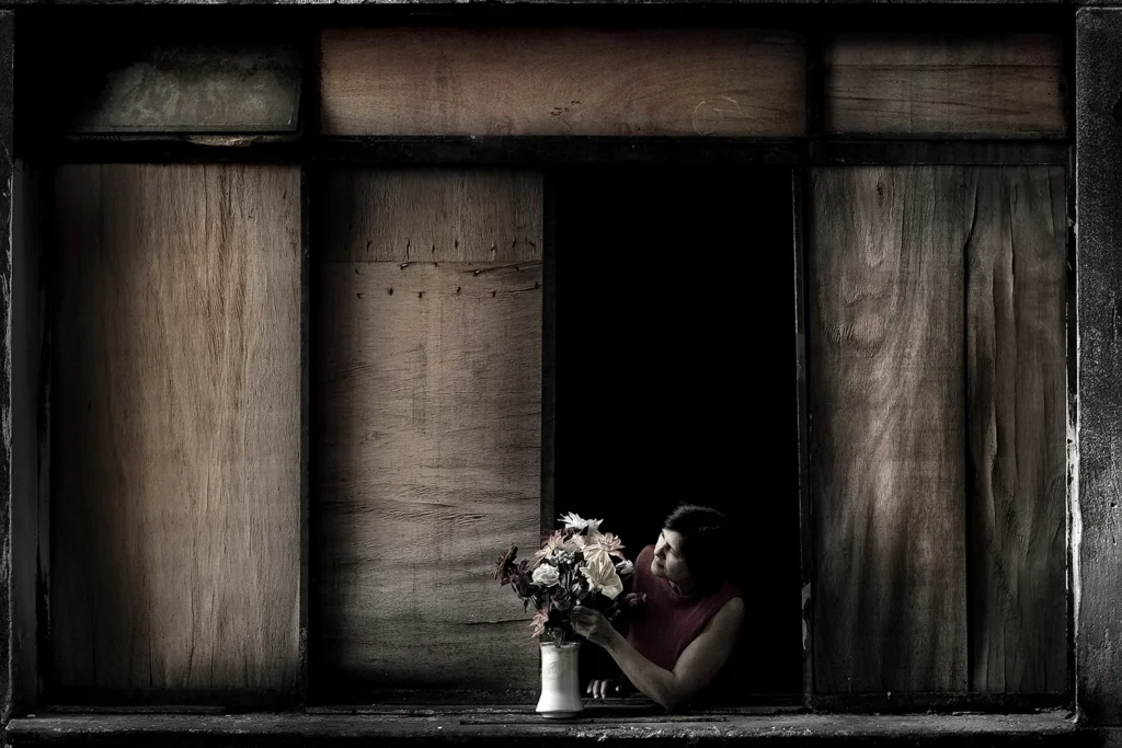 Fotografía de Julio Bittencourt que muestra a una persona en un interior oscuro arreglando flores junto a una ventana de madera.