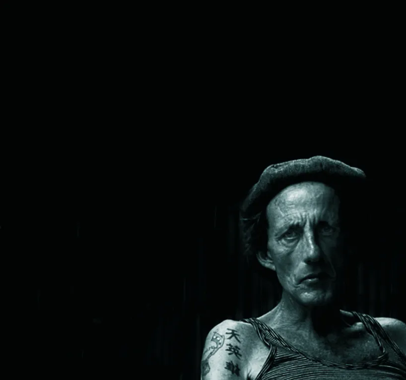 Retrato en primer plano de una persona con rostro marcado, tatuaje en el brazo y fondo oscuro, iluminado con contraste dramático.