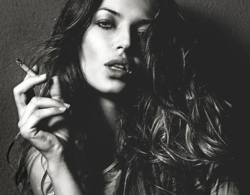 Retrato en blanco y negro de una persona con cabello largo y ondulado, sosteniendo un cigarrillo, con expresión relajada y mirada directa a cámara.