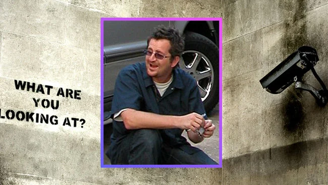 Hombre posando frente a una cámara de vigilancia con el mensaje “What are you looking at?” en una pared.