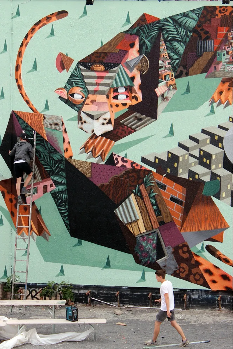 Mural en proceso: un tigre geométrico y texturizado es pintado sobre una fachada verde, con árboles triangulares y ciudad en el fondo.