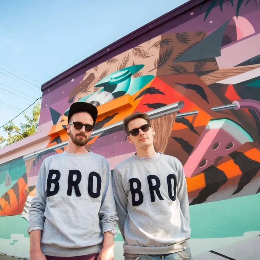 Dos personas con sudaderas grises que dicen "BRO" posan frente a un mural vibrante de Low Bros con formas abstractas, frutas estilizadas y colores intensos.