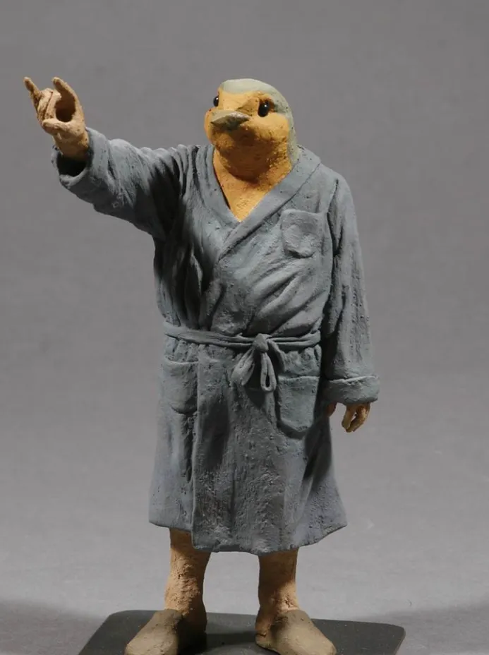 Figura antropomórfica con cabeza de ave y cuerpo humano vestido con bata gris, haciendo gesto con la mano sobre base negra.