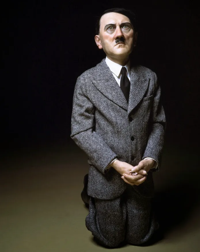 Escultura hiperrealista de figura masculina arrodillada con traje gris, manos juntas y rasgos faciales que evocan a Adolf Hitler, sobre fondo oscuro.
