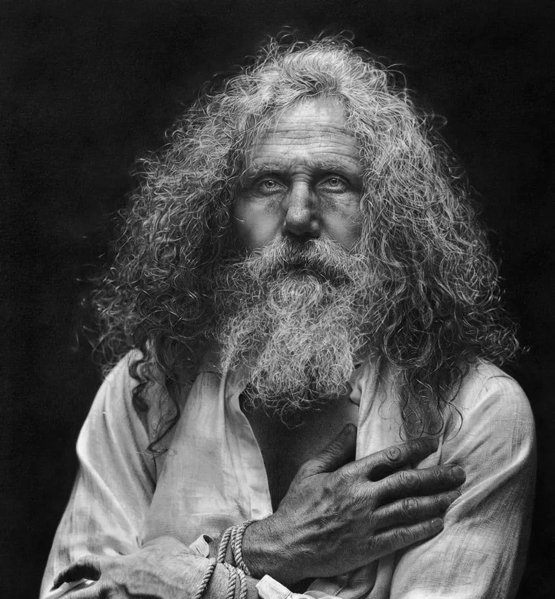 Retrato hiperrealista en blanco y negro de un hombre mayor con barba y cabello largo, iluminado dramáticamente sobre fondo oscuro.