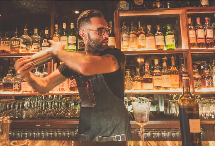 Bartender preparando cóctel en barra bien surtida, con fondo de botellas y copas colgantes, en ambiente cálido y acogedor.