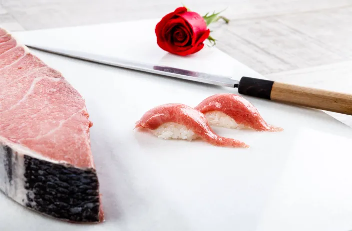 Composición culinaria con corte de atún crudo, nigiri sushi, cuchillo japonés y rosa roja sobre fondo claro de madera.