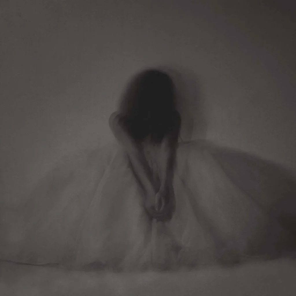 Figura femenina sentada con cabeza inclinada y vestido claro en entorno oscuro y difuso