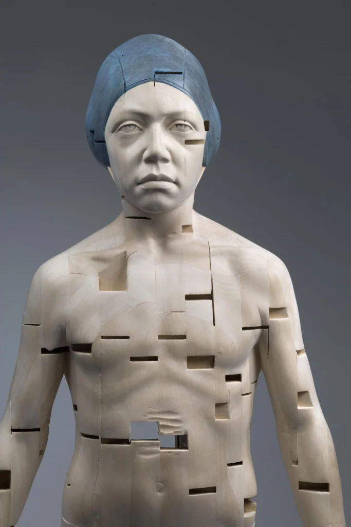 Escultura de torso humano en madera con gorra azul, marcada por cortes geométricos que fragmentan la figura.