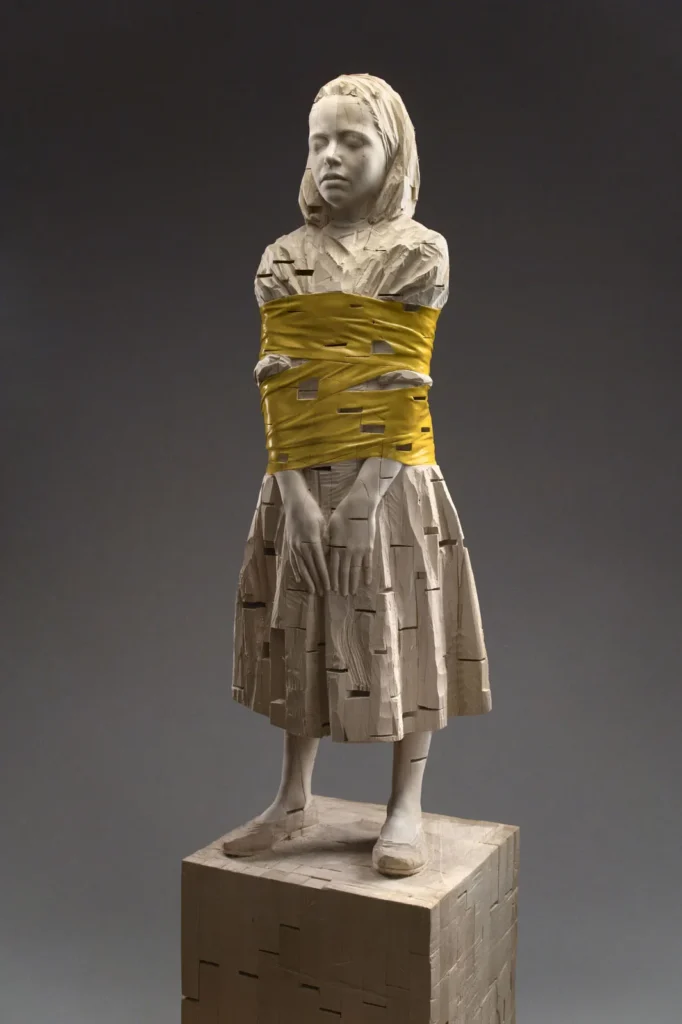 Escultura de madera de una niña con el torso envuelto en cinta amarilla, obra contemporánea de Gehard Demetz.