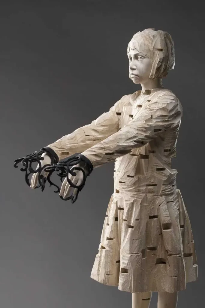 Escultura de madera de una niña con detalles tallados geométricamente y manos adornadas con estructuras negras, obra contemporánea de Gehard Demetz.