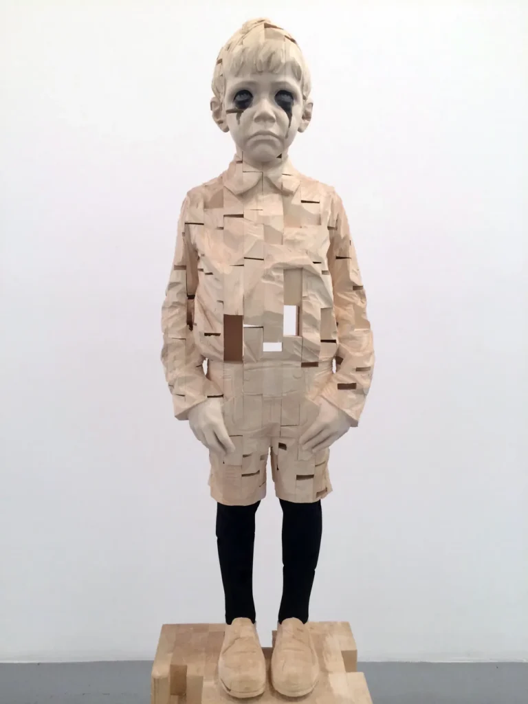 Escultura de torso humano en madera con gorra azul, marcada por cortes geométricos que fragmentan la figura.