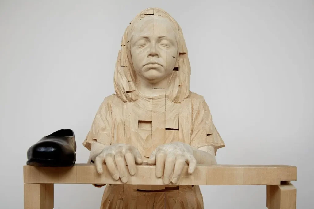 Escultura de madera de figura humana con ojos cerrados, compuesta por segmentos geométricos, sentada frente a una mesa con un zapato negro.