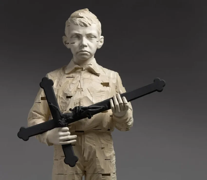 Escultura de niño con rostro pálido y cuerpo tallado en madera, sosteniendo una cruz negra estilizada con expresión solemne.