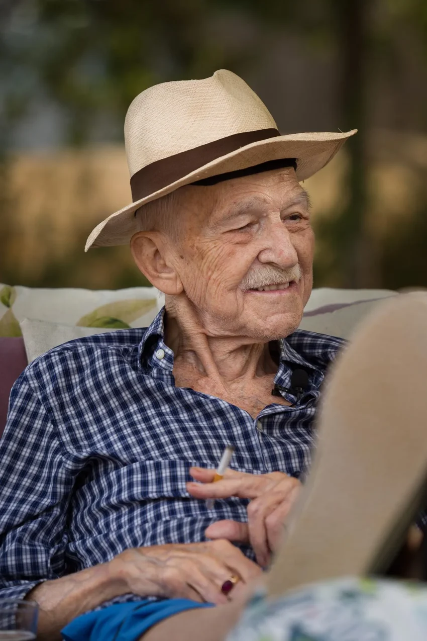 Antonio Escohotado sentado al aire libre con sombrero de paja, camisa de cuadros y cigarrillo en mano.
