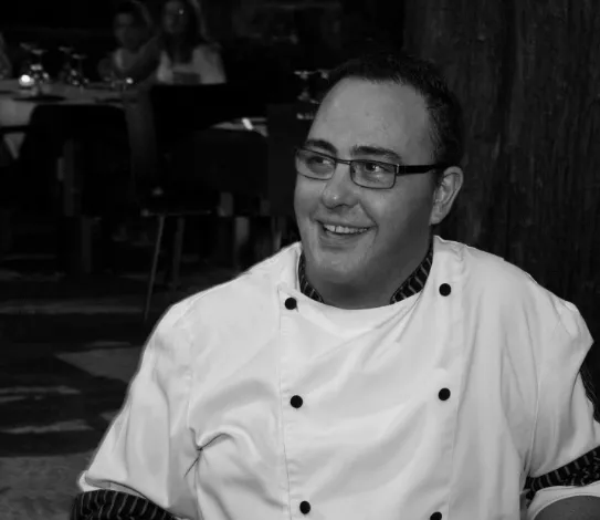Chef Dario Zappala sonriente con gafas y chaqueta de cocina en La Búfala de Alaró, con comensales al fondo en ambiente de restaurante.
