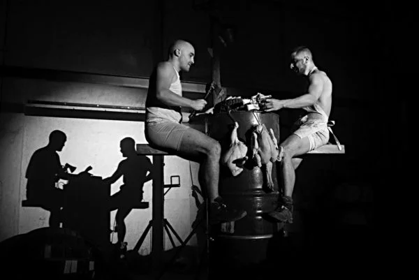 Dos intérpretes de La Fura dels Baus manipulan piezas de carne colgadas mientras sus sombras proyectan una escena que recuerda a dos personas sentadas a una mesa, en una imagen de la obra Manes.