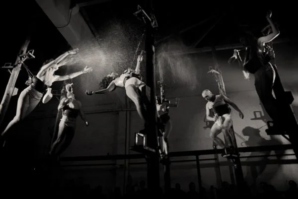 Intérpretes de La Fura dels Baus suspendidos en el aire durante una escena acrobática de la obra Manes, iluminados dramáticamente mientras realizan movimientos físicos intensos.