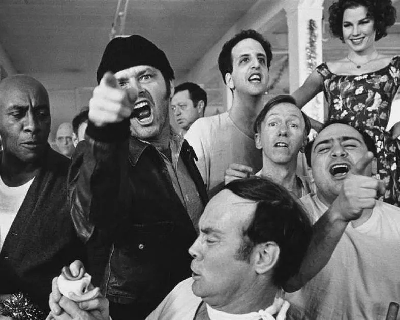 Escena en blanco y negro de un grupo de personas en un centro institucional, con un hombre al frente gritando y señalando mientras otros reaccionan a su alrededor.