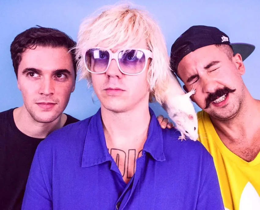 Tres miembros de la banda londinense Dancehall posan frente a un fondo azul; uno lleva gafas moradas y sostiene una rata blanca que se acerca al rostro de otro integrante.