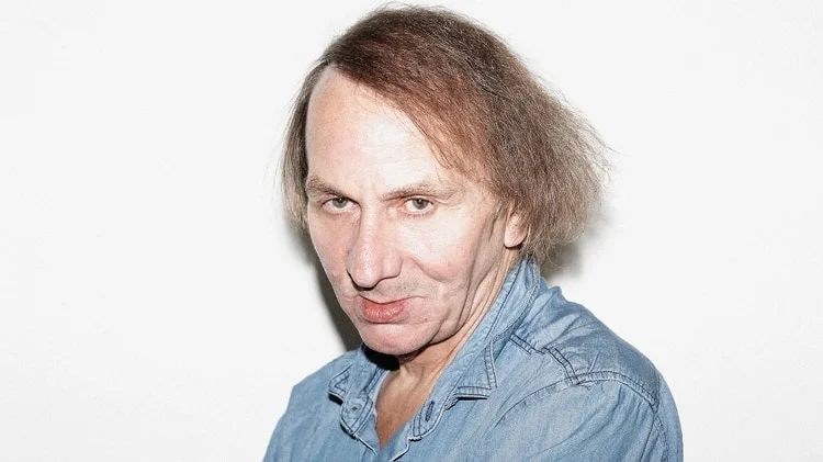 Retrato de Michel Houellebecq con camisa vaquera azul sobre fondo blanco.