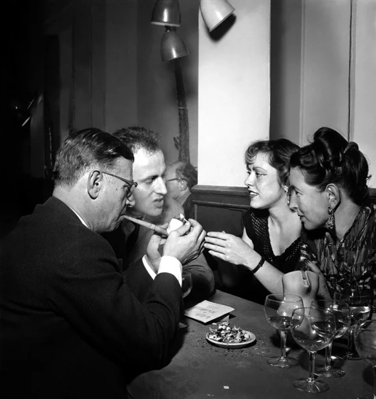 Grupo de intelectuales conversando y fumando en una mesa de café con copas de vino en una fotografía en blanco y negro de mediados del siglo XX.