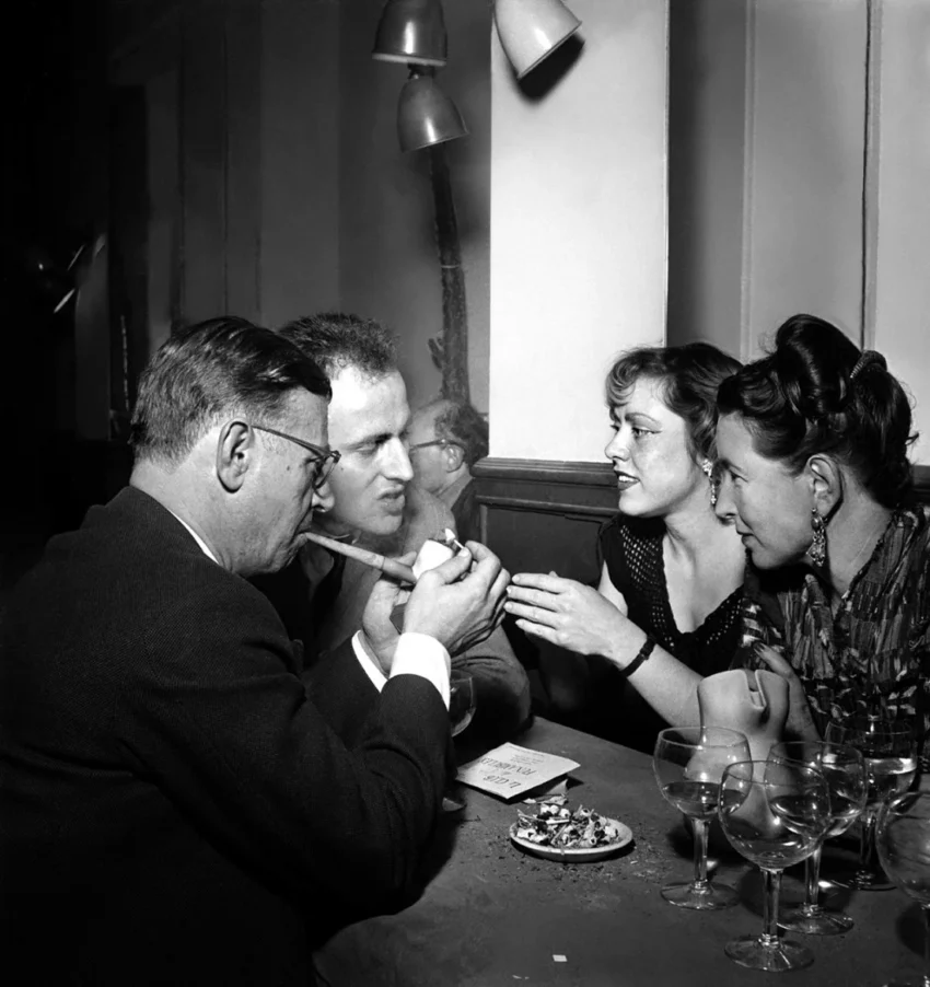 Grupo de intelectuales conversando y fumando en una mesa de café con copas de vino en una fotografía en blanco y negro de mediados del siglo XX.