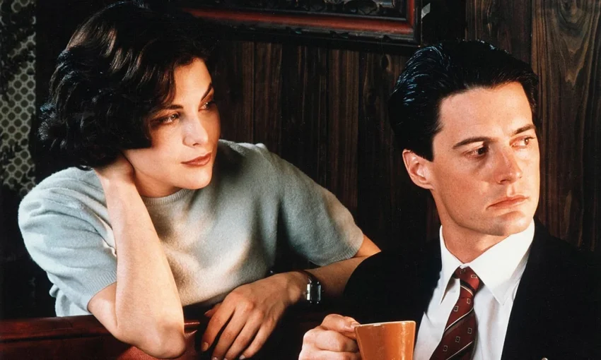 Dos personas sentadas en un café de madera: una con camisa gris, otra con traje oscuro y taza naranja.