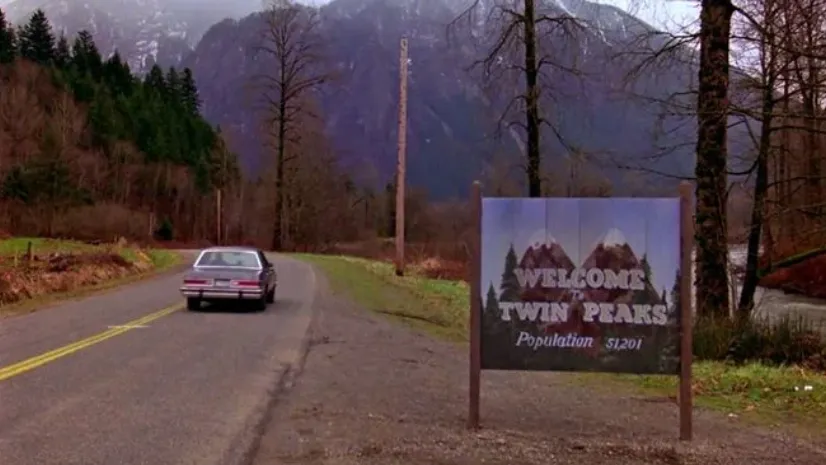 Carretera rural entre montañas con cartel de bienvenida a Twin Peaks y coche alejándose.