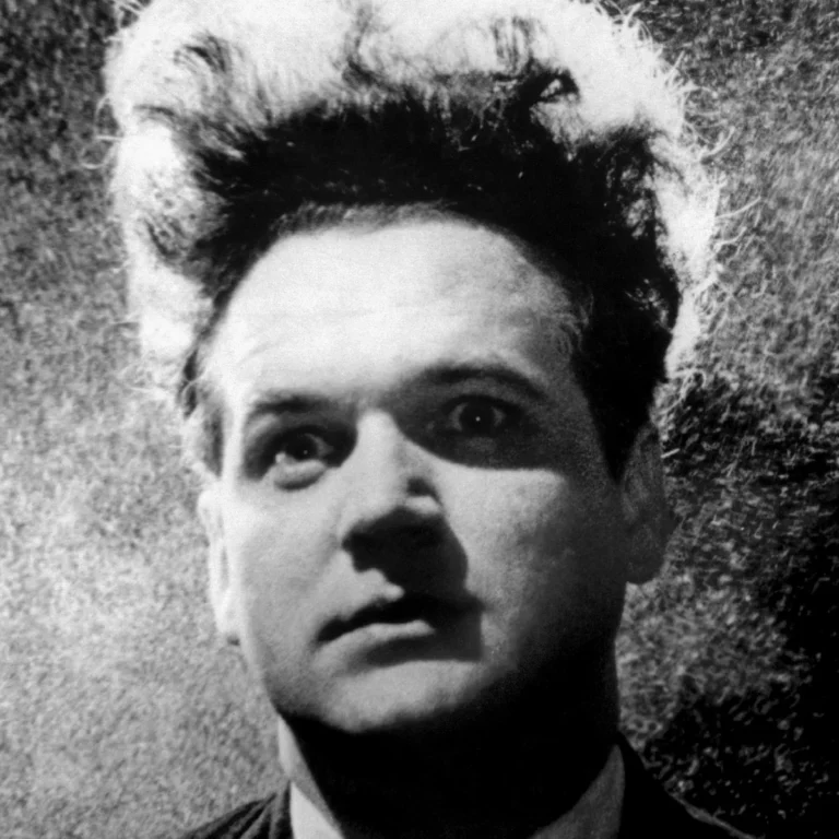 David Lynch en blanco y negro, de pie en ascensor, bajo luz dramática, en escena surrealista.