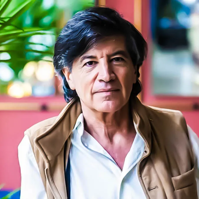 Carlos López-Otín con camisa blanca y chaleco beige, retrato en interior con fondo rojo y planta decorativa.