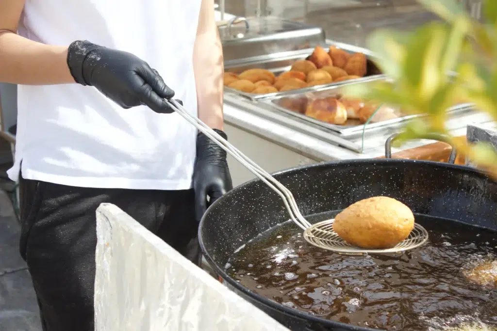 Persona con camisa blanca y guantes negros levantando alimento frito dorado con espumadera metálica desde olla negra con aceite; cocina exterior con bandeja de frituras al fondo.