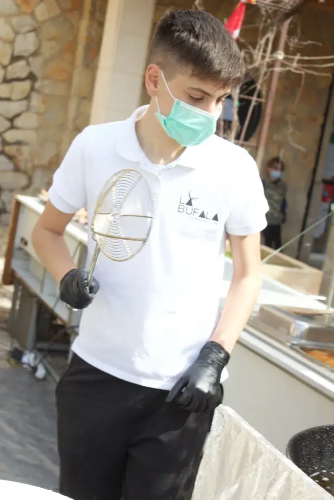 Persona joven con polo blanco “BUFALA”, guantes negros y mascarilla verde, sosteniendo espumadera metálica en cocina exterior con muros de piedra.