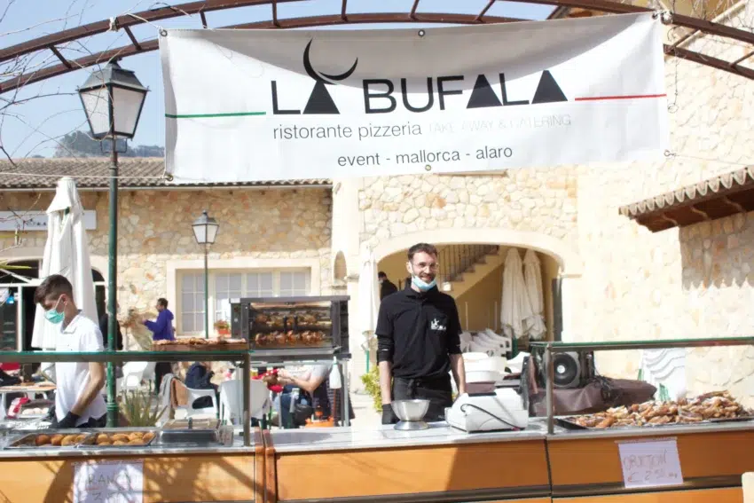 Puesto de comida exterior frente a edificio de piedra con arcos; banner de “LA BUFALA” anuncia restaurante, pizzería, take away y catering; dos personas atienden el mostrador con pollos asados y repostería.
