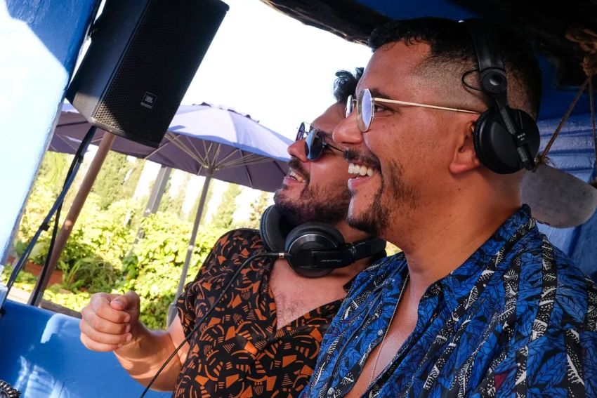 Dos DJs mezclando al aire libre junto a un altavoz, rodeados de vegetación y luz natural.