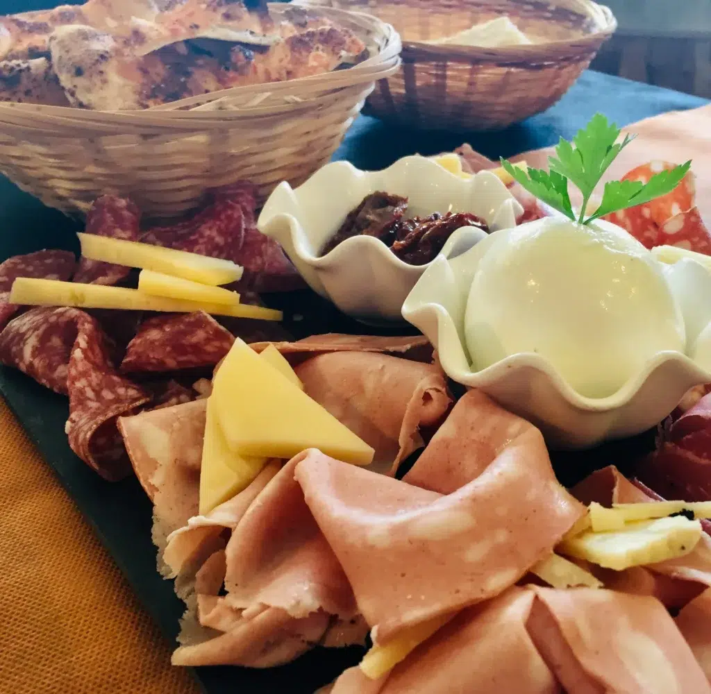 Tabla de embutidos y quesos con salami, mortadela, quesos amarillos y mozzarella decorada con perejil; acompañada de tomates secos y pan en cestas de mimbre.