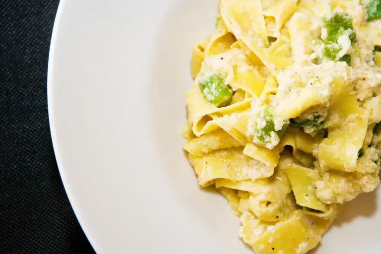 Plato de pasta tipo pappardelle con salsa blanca cremosa, ricotta y verduras verdes sobre superficie oscura.