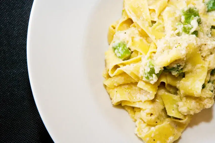 Plato de pasta tipo pappardelle con salsa blanca cremosa, ricotta y verduras verdes sobre superficie oscura.