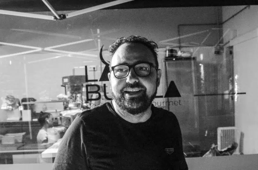 Sario Zappala con gafas y barba en cocina gourmet, de pie frente a cristal con el texto “LA Bu Gourmet”; fondo con equipo culinario y figura trabajando.
