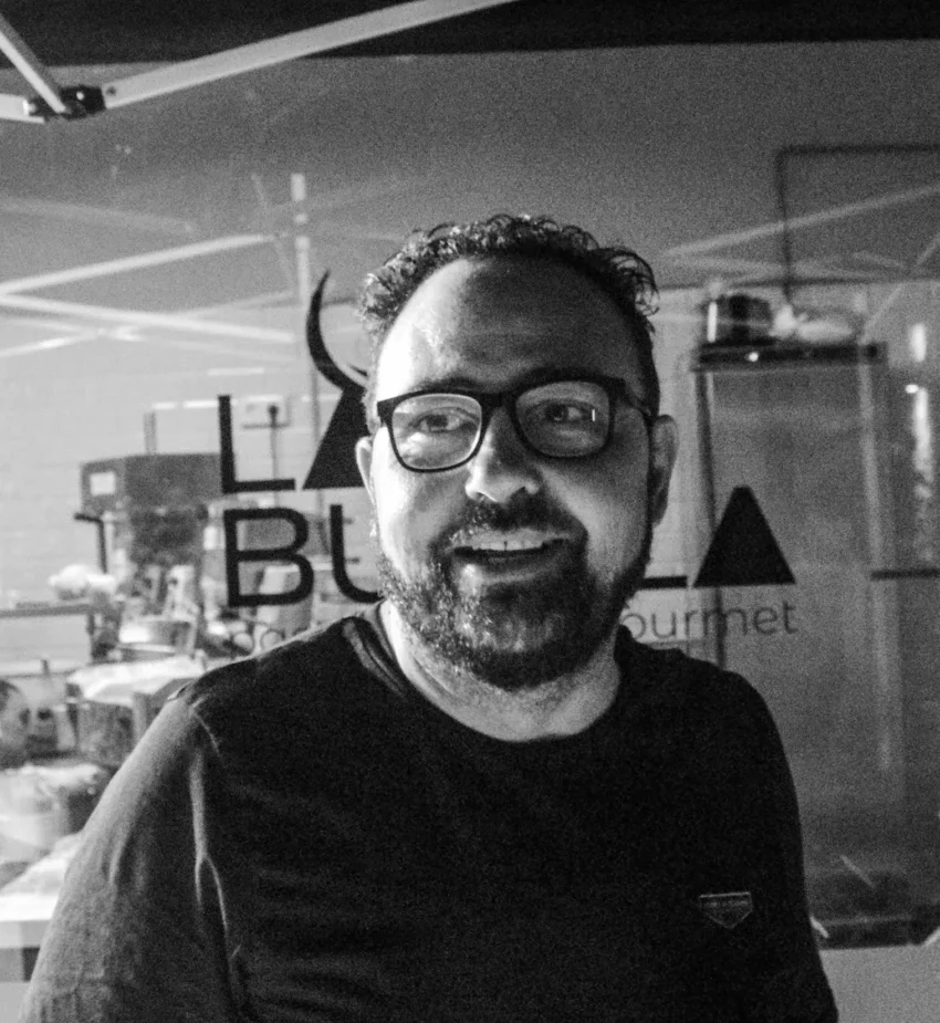 Dario Zappala con gafas y barba en cocina gourmet, de pie frente a cristal con el texto “LA Bu Gourmet”; fondo con equipo culinario y figura trabajando.