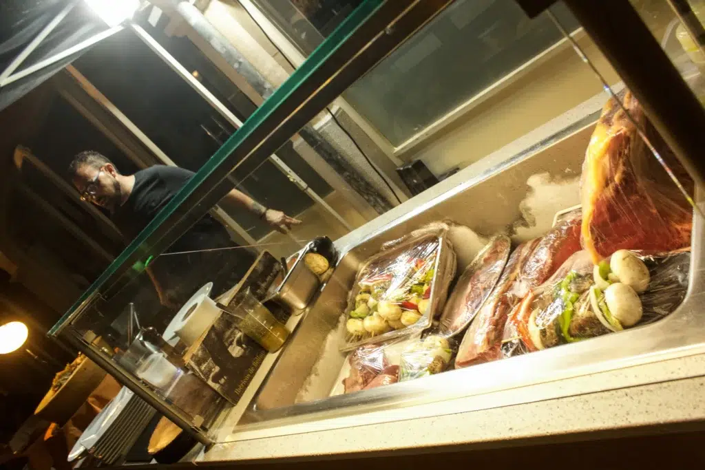 Mostrador de alimentos con bandejas de carne cruda y verduras envueltas en plástico; jamón curado en posición vertical; ambiente de cocina profesional.