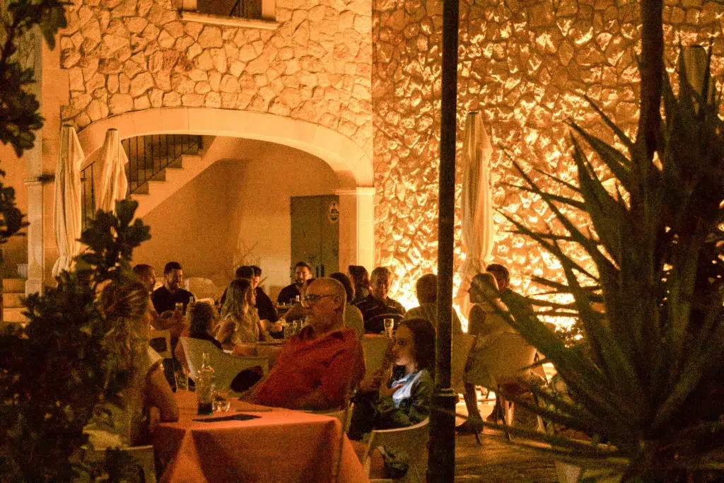 Terraza nocturna con mesas cubiertas de manteles naranjas, comensales conversando, iluminación cálida, plantas decorativas y arquitectura rústica mediterránea.