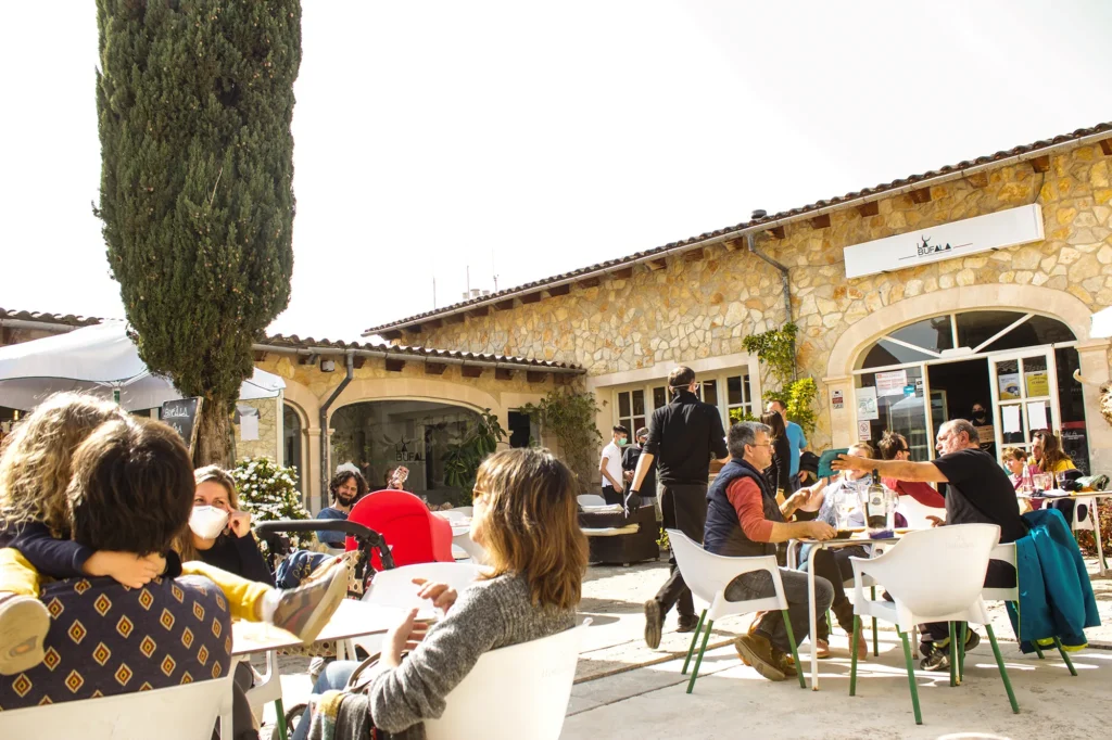 Comensales en terraza de La Búfala de Alaró, disfrutando de comida y conversación frente a fachada de piedra bajo luz natural.