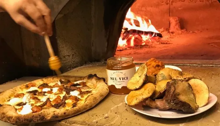 Pizza recién horneada con ricotta y embutido picante, rociada con miel de azahar frente a horno de leña encendido.