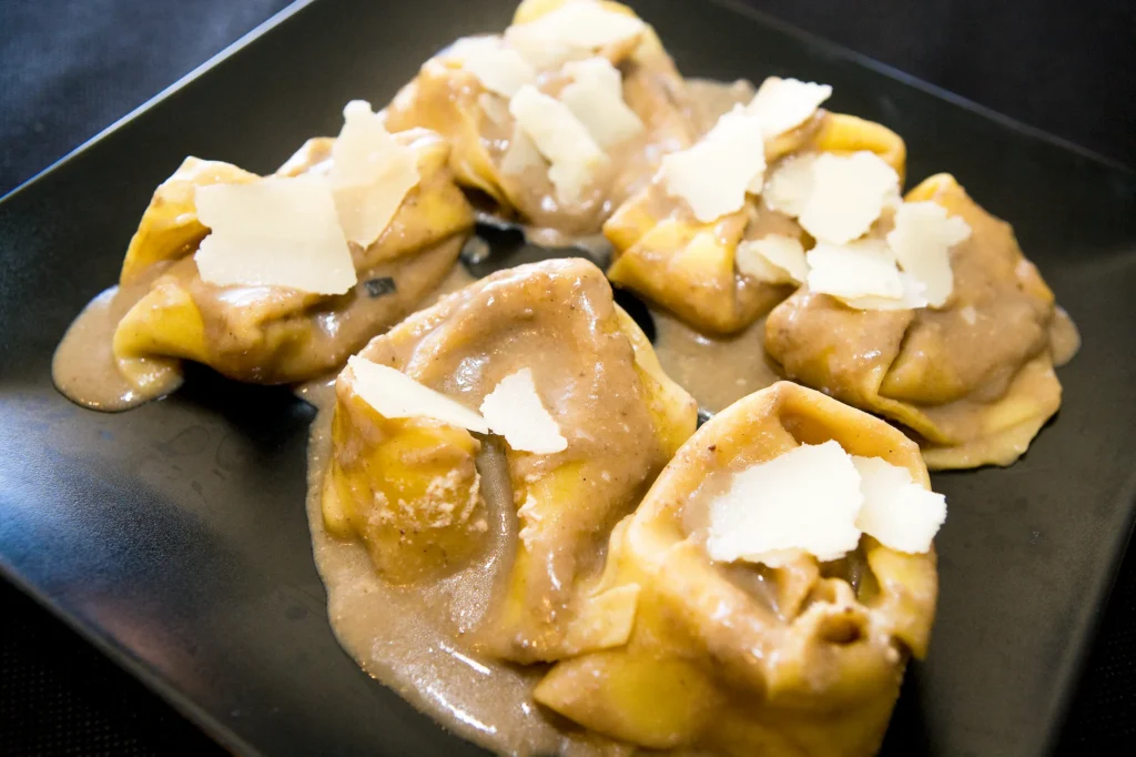 Tortellini en salsa cremosa marrón con virutas de parmesano sobre plato negro cuadrado.