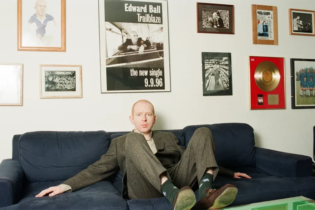 Alan McGee sentado en un sofá azul, rodeado de memorabilia musical, en una escena íntima y vintage.