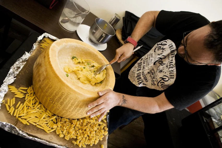 Dario de La Búfala de Alaró mezclando pasta con salsa cremosa dentro de una rueda de queso Parmigiano-Reggiano; mesa con variedades de pasta cruda, utensilios y decoración culinaria.