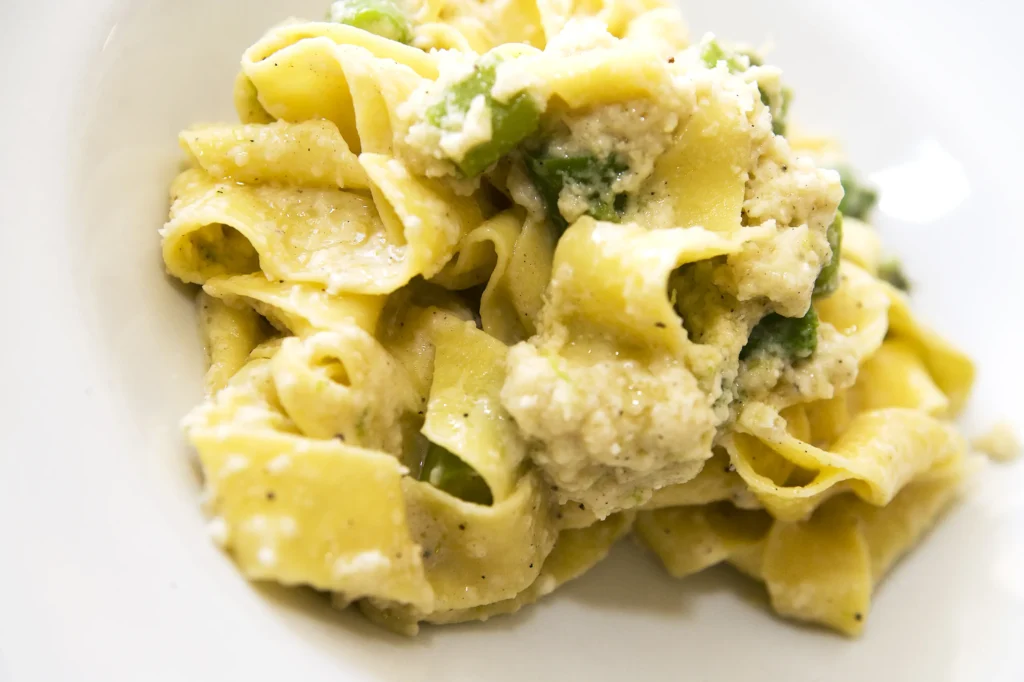 Plato de pasta tipo pappardelle con salsa blanca cremosa, ricotta y verduras verdes; granulado visible y pimienta negra espolvoreada sobre superficie blanca.
