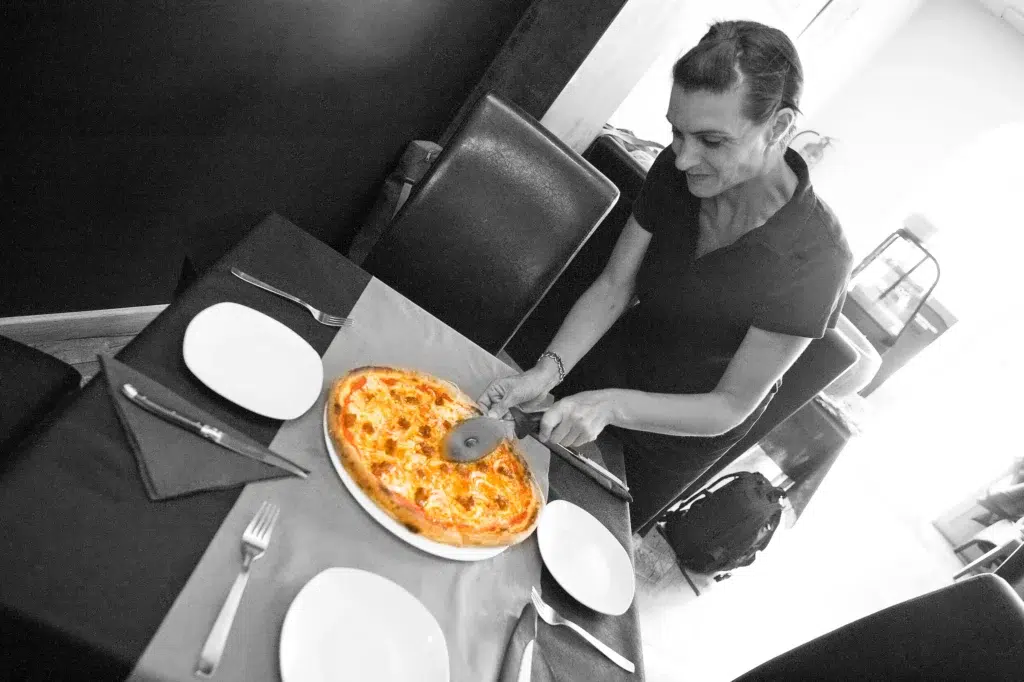 Yara cortando pizza con rueda metálica sobre plato blanco en mesa preparada con cubiertos y servilletas negras; ambiente interior con sillas negras y fondo oscuro.