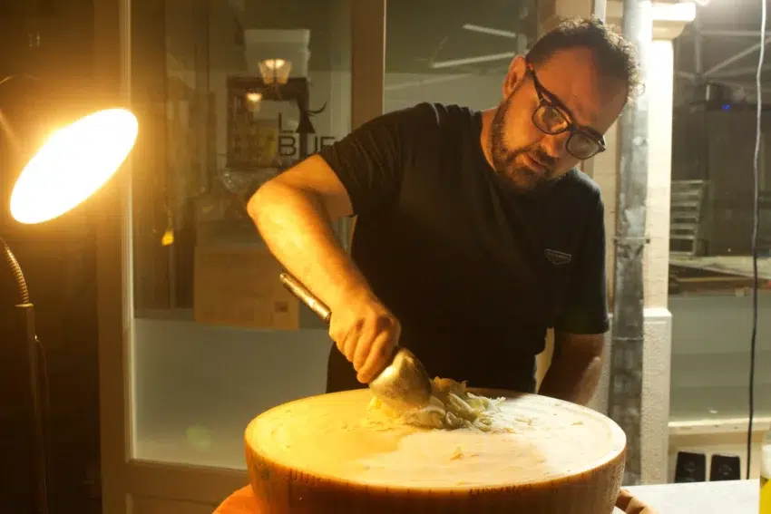 Dario Zappalà rallando queso desde una rueda grande sobre base de madera, bajo luz cálida; ambiente interior con reflejos en cristal y atmósfera concentrada.